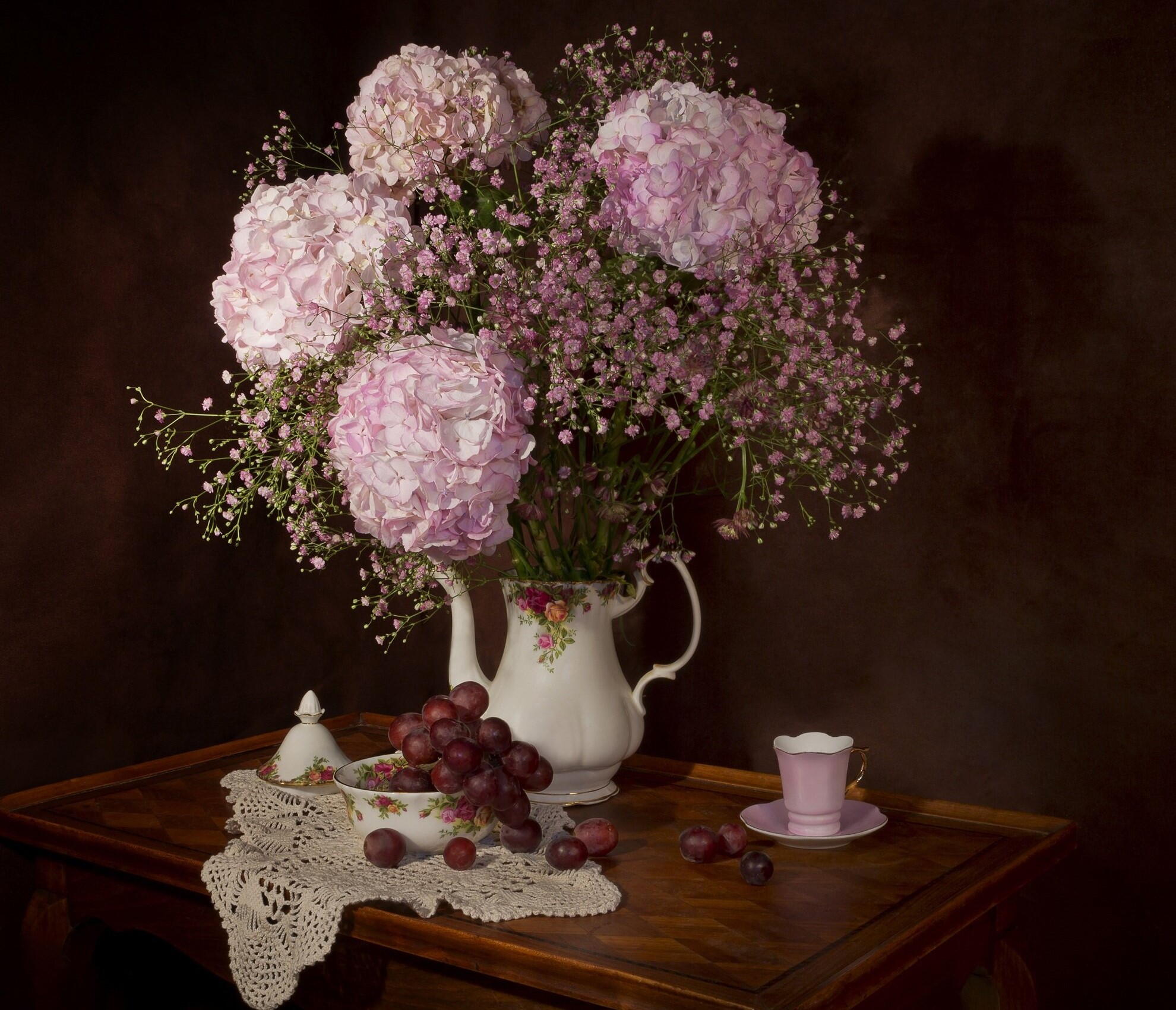 Bule utilizado como vaso, repleto de gypsophilas e outras flores. Ao redor, uvas e xícaras completam a decoração